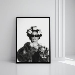 May include: Black and white framed print of a person wearing sunglasses, a fur coat, and a money-themed hair accessory, holding a cigarette and a cocktail glass. The image is displayed against a white wall.