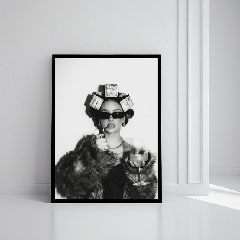 May include: Black and white framed print of a person wearing sunglasses, a fur coat, and a money-themed hair accessory, holding a cigarette and a cocktail glass. The image is displayed against a white wall.
