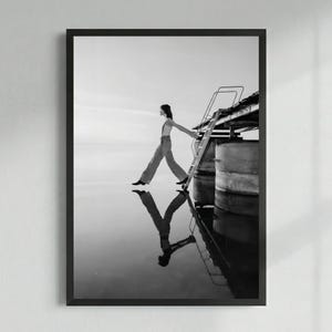 Puede incluir: Impresión artística enmarcada en blanco y negro. Una persona camina sobre una superficie reflectante, con una escalera y una estructura de muelle en el fondo. La imagen es una fotografía artística de alto contraste.