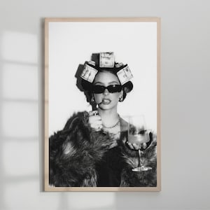 May include: Black and white framed print of a woman wearing sunglasses, a fur coat, and money curlers in her hair. She holds a glass of a drink and a cigarette. The print is in a wooden frame.