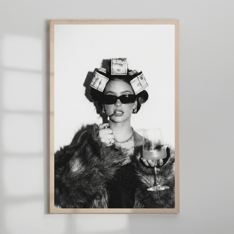 May include: Black and white framed print of a woman wearing sunglasses, a fur coat, and money curlers in her hair. She holds a glass of a drink and a cigarette. The print is in a wooden frame.