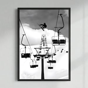 May include: A black and white framed wall art print featuring a snowboarder mid-air performing a jump above a ski lift system. The composition shows multiple empty chairlifts suspended by cables against a snowy mountain backdrop and cloudy sky.