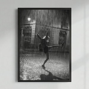 May include: Black and white framed print of a person dancing in the rain. The person is silhouetted against a city backdrop, with arms outstretched and one leg extended. The rain is heavy, creating a dramatic effect.