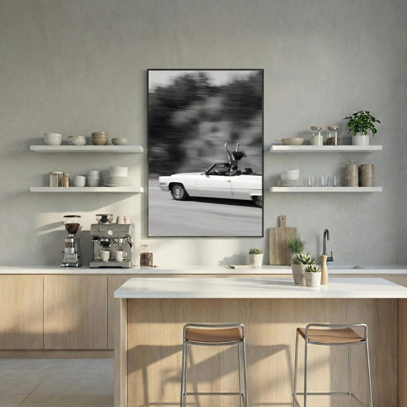 May include: Monochrome framed print showing a white convertible car in motion, with a person standing up in the car with their arms raised. The background is blurred, suggesting speed. The print is displayed in a modern kitchen setting.