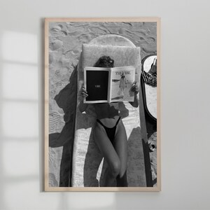 Peut inclure: Tirage encadr&eacute; en noir et blanc d'une personne lisant un magazine sur une chaise longue de plage. Le magazine ouvert affiche le texte "THE VIBE" et "VIBE". La personne porte des lunettes de soleil et un maillot de bain noir.