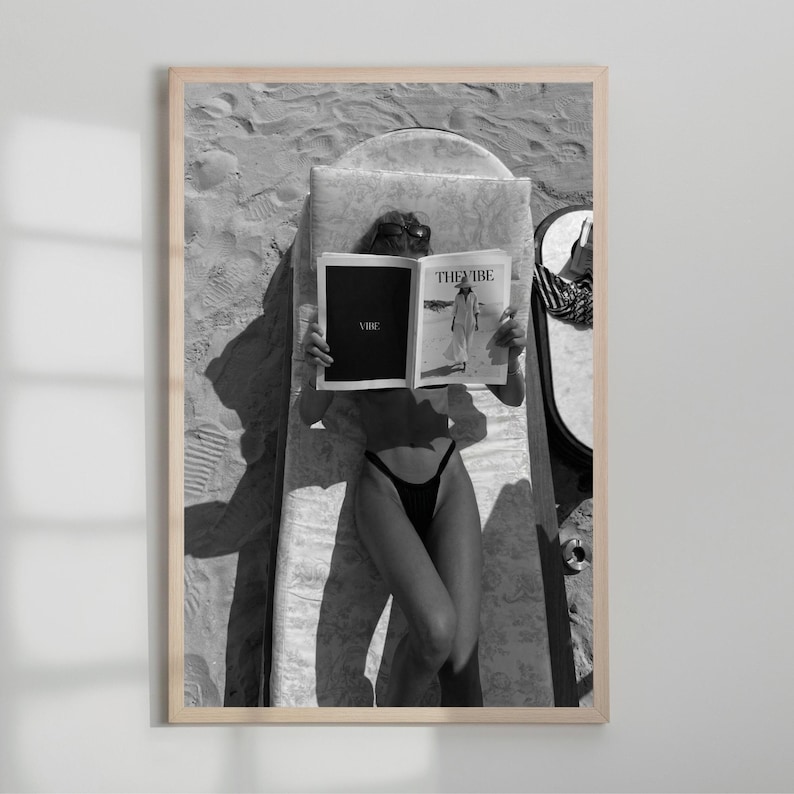 Peut inclure: Tirage encadr&eacute; en noir et blanc d'une personne lisant un magazine sur une chaise longue de plage. Le magazine ouvert affiche le texte "THE VIBE" et "VIBE". La personne porte des lunettes de soleil et un maillot de bain noir.