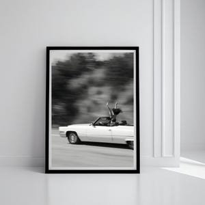 May include: Monochrome framed print showing a white vintage convertible car in motion on a road. A person is standing in the car with arms raised. The background is blurred, indicating speed and movement.