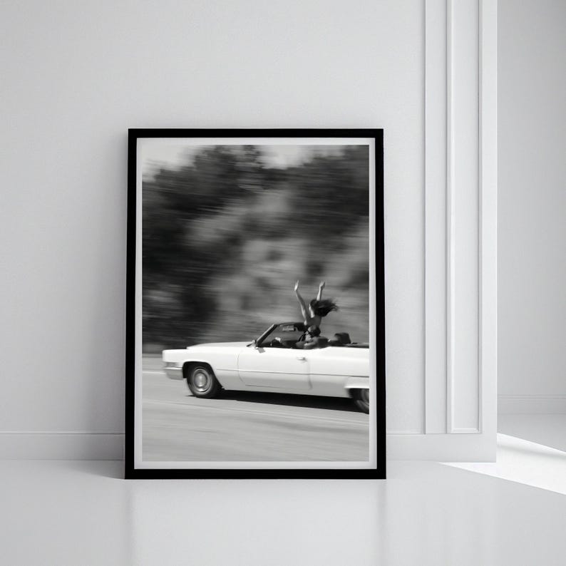 May include: Monochrome framed print showing a white vintage convertible car in motion on a road. A person is standing in the car with arms raised. The background is blurred, indicating speed and movement.