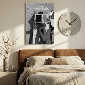 Peut inclure: Photographie en noir et blanc d'une personne lisant un magazine sur une chaise longue. L'image est un gros plan, avec la personne et le magazine occupant la majeure partie du cadre. Une grande horloge ronde est sur le mur. Un lit est au premier plan.