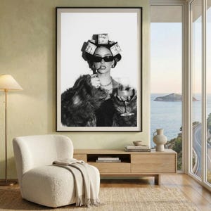 May include: Black and white framed print of a person wearing sunglasses, a fur coat, and hair rollers made of money, holding a cigarette and a glass of wine. The print is displayed above a wooden cabinet and a white chair.