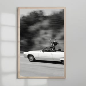 May include: Monochrome framed print featuring a white convertible car in motion. A person is standing in the car with their arms raised. The background is blurred, implying speed. The image conveys a feeling of liberation and adventure.