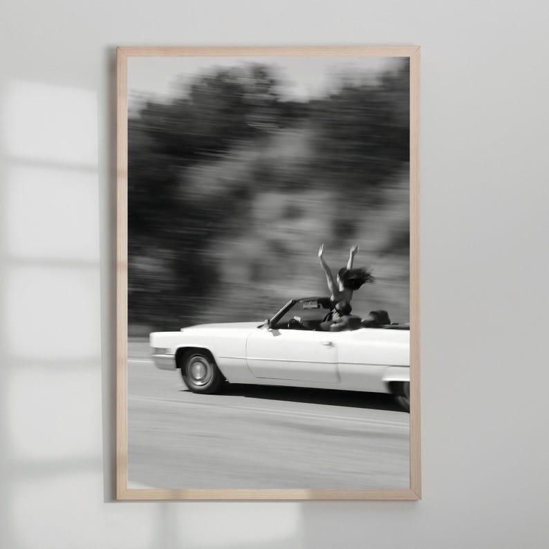 May include: Monochrome framed print featuring a white convertible car in motion. A person is standing in the car with their arms raised. The background is blurred, implying speed. The image conveys a feeling of liberation and adventure.