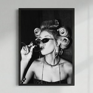 May include: Monochrome framed print featuring a person wearing sunglasses and hair curlers, drinking wine. The person is also wearing a necklace and earrings. The image has a vintage, glamorous aesthetic.
