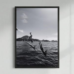 May include: Black and white framed print featuring three figures diving into the ocean. The image captures the moment of the divers' descent, with the sea and a mountainous landscape in the background under a bright sky.