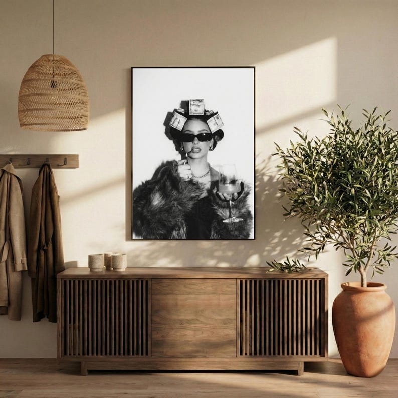 May include: Black and white framed print of a woman wearing sunglasses, a fur coat, and hair rollers made of money, holding a cigarette and a wine glass. The print is displayed above a wooden cabinet with vertical slats.