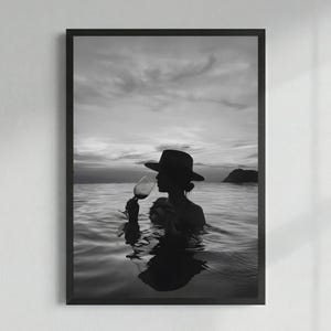 May include: Black and white framed print of a person in a hat drinking wine in the ocean. The silhouette of the person is visible against the water and sky. The image is framed in a black border.