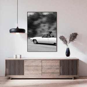 May include: Monochrome framed print showing a white vintage convertible car in motion. A person is standing in the car with arms raised. The background is blurred, indicating speed. The print is above a wooden cabinet.