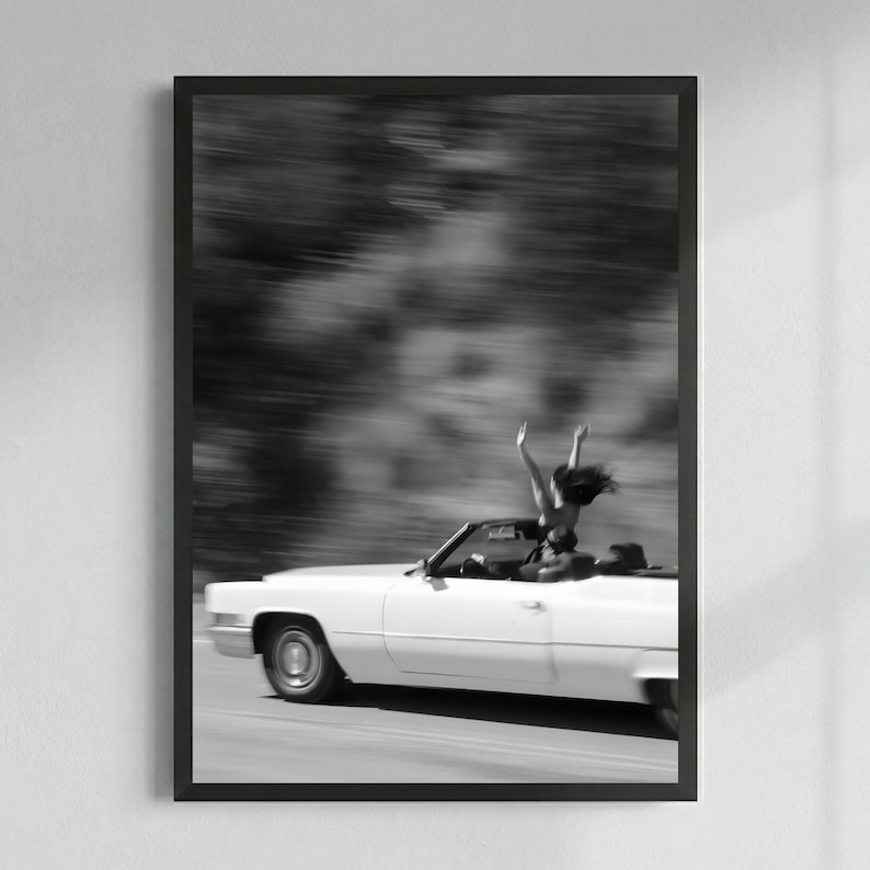 May include: Monochrome framed print featuring a white convertible car speeding along a road. A person stands in the car with arms raised. The background is blurred, conveying motion and speed.