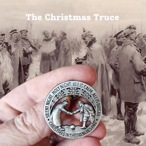 May include: A silver-coloured commemorative coin featuring the text "Silent Night, Holy Night, All is Calm, All is Bright" and "Christmas in the Trenches 1914". The coin shows two soldiers shaking hands. The background is a sepia-toned image of soldiers.