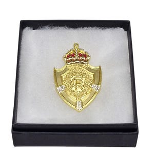 Puede incluir: Un broche dorado en forma de escudo con una corona y la letra "R" en el centro, presentado en una caja negra con relleno blanco. La corona tiene detalles rojos y el escudo adornos decorativos.