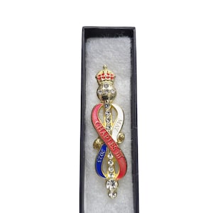 Può includere: Una spilla dorata con una corona, un nastro con la scritta "CHARLES III MAY 2023" e dettagli in cristallo trasparente. La spilla è in una scatola nera con imbottitura bianca.