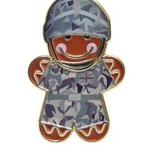 Puede incluir: Un pin de hombre de jengibre con forma de soldado, con uniforme y casco de camuflaje. El pin tiene un contorno dorado y presenta la bandera de la Union Jack en el uniforme. El hombre de jengibre tiene una cara sonriente con mejillas rosadas.