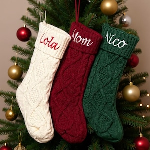 May include: Three cable-knit Christmas stockings hang on a decorated tree. One is cream-coloured with "Lola" embroidered in red, another is burgundy with "Mom" in white, and the third is green with "Nico" in white.