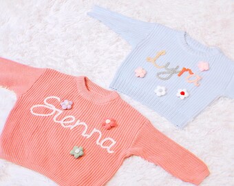 May include: Two knitted baby sweaters with embroidered names, Sienna and Lyra, and flower embellishments. The sweater on the left is pink and the sweater on the right is light blue.
