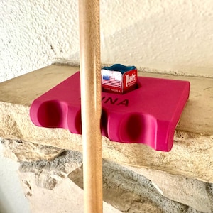 May include: A hot pink billiard cue holder with three circular cue slots. A small box of Master billiard chalk with an American flag design sits on top. A wooden cue is in the foreground.