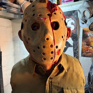 May include: A tan hockey mask with numerous small holes, bloodstains, and a split down the middle. The mask is worn over a tan shirt with a buttoned pocket. The image is a bust of a horror movie character.