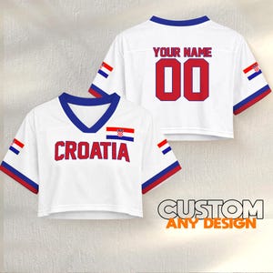 K&ouml;nnte beinhalten: Wei&szlig;es Crop-Top-Trikot mit "CROATIA" in roten Buchstaben und der kroatischen Flagge. Auf der R&uuml;ckseite steht "YOUR NAME" &uuml;ber der Nummer "00" in Rot. Blaue und rote Akzente an &Auml;rmeln und Ausschnitt. "CUSTOM ANY DESIGN" ist unten aufgedruckt.