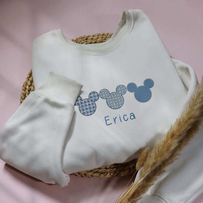 May include: White sweatshirt with three blue embroidered Mickey Mouse heads and the name "Erica" embroidered below.