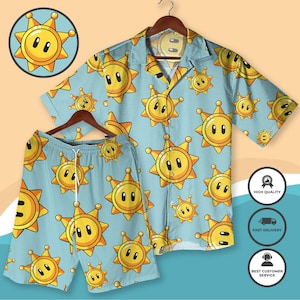 Puede incluir: Conjunto de camisa de manga corta y pantalón corto azul claro. El conjunto presenta un patrón repetido de personajes amarillos con forma de sol y caras sonrientes. La camisa está colgada en una percha de madera.