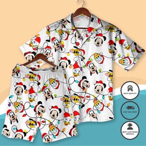 Puede incluir: Conjunto de pijama blanco con temática navideña, con una camisa de manga corta con botones y pantalones cortos a juego. El conjunto está adornado con un patrón repetido de personajes de Mickey Mouse, Pato Donald y Goofy con gorros de Papá Noel y luces navideñas.