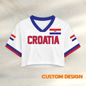 K&ouml;nnte beinhalten: Wei&szlig;es Crop-Jersey mit dem Wort "CROATIA" in roten Blockbuchstaben. Das Trikot hat einen blauen V-Ausschnitt und rot-blaue Verzierungen an den &Auml;rmeln. Die kroatische Flagge ist auf der Brust und den &Auml;rmeln abgebildet. Der Text "CUSTOM DESIGN" steht unten.