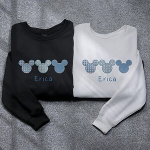 May include: Two long-sleeved sweatshirts with a row of three blue and white Mickey Mouse ears embroidered on each. The text "Erica" is embroidered below the ears on each sweatshirt. One sweatshirt is black and the other is white.