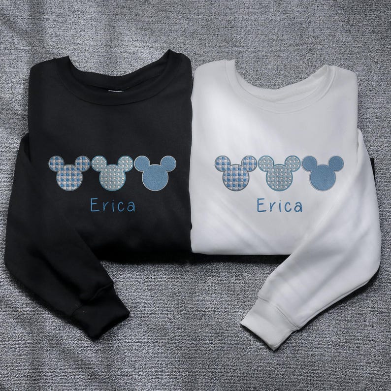 May include: Two long-sleeved sweatshirts with a row of three blue and white Mickey Mouse ears embroidered on each. The text "Erica" is embroidered below the ears on each sweatshirt. One sweatshirt is black and the other is white.