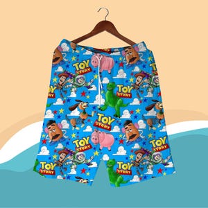 K&ouml;nnte beinhalten: Blaue Shorts mit einem sich wiederholenden Muster von Toy Story-Charakteren, darunter Woody, Buzz Lightyear und Mr. Potato Head. Die Shorts haben einen Kordelzug in der Taille und h&auml;ngen an einem Holzb&uuml;gel.