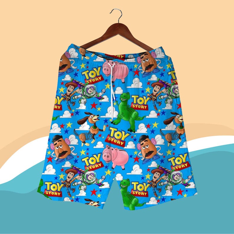 K&ouml;nnte beinhalten: Blaue Shorts mit einem sich wiederholenden Muster von Toy Story-Charakteren, darunter Woody, Buzz Lightyear und Mr. Potato Head. Die Shorts haben einen Kordelzug in der Taille und h&auml;ngen an einem Holzb&uuml;gel.