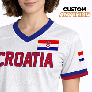 K&ouml;nnte beinhalten: Wei&szlig;es Trikot mit blauem V-Ausschnitt und rot-blauen &Auml;rmelbesatz. Das Wort "CROATIA" ist rot &uuml;ber die Brust gedruckt. Eine kroatische Flagge ist auf Brust und &Auml;rmel abgebildet. Der Text "CUSTOM ANYTHING" befindet sich oben rechts.
