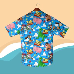 K&ouml;nnte beinhalten: Ein kurz&auml;rmeliges Hemd mit einem farbenfrohen Muster von Figuren aus dem Animationsfilm "Toy Story". Das Design zeigt Woody, Buzz Lightyear, Rex, Hamm und Mr. Potato Head auf blauem Hintergrund mit Wolken, Sternen und den Worten "Toy Story".