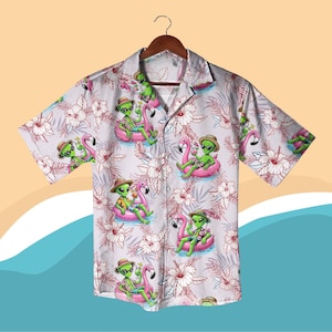 Puede incluir: Una camisa de manga corta con fondo gris claro y estampado floral. La camisa presenta un diseño repetido de extraterrestres verdes con sombreros y gafas de sol, flotando en flotadores de piscina de flamencos rosas. La camisa tiene un cuello clásico y un corte relajado.