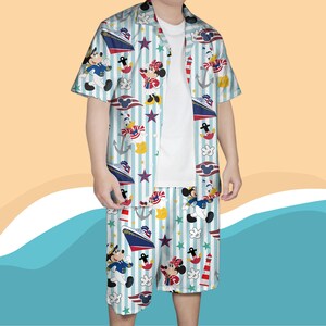 May include: A two-piece matching set featuring a short-sleeved button-down shirt and shorts. The set is light blue with vertical white stripes and a nautical-themed print of cartoon characters, ships, anchors, and stars. The shirt is open, revealing a white t-shirt underneath.