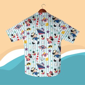 May include: A short-sleeved button-down shirt with a nautical theme. The shirt features a pattern of Mickey and Minnie Mouse, Donald Duck, anchors, lighthouses, and cruise ships on a light blue and white striped background. The shirt is hanging on a wooden hanger.