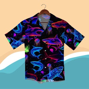 May include: Black button-down shirt featuring a neon shark and jellyfish design. The shirt showcases sharks in blue, pink, and orange, and jellyfish in green and purple. The shirt is displayed on a wooden hanger.