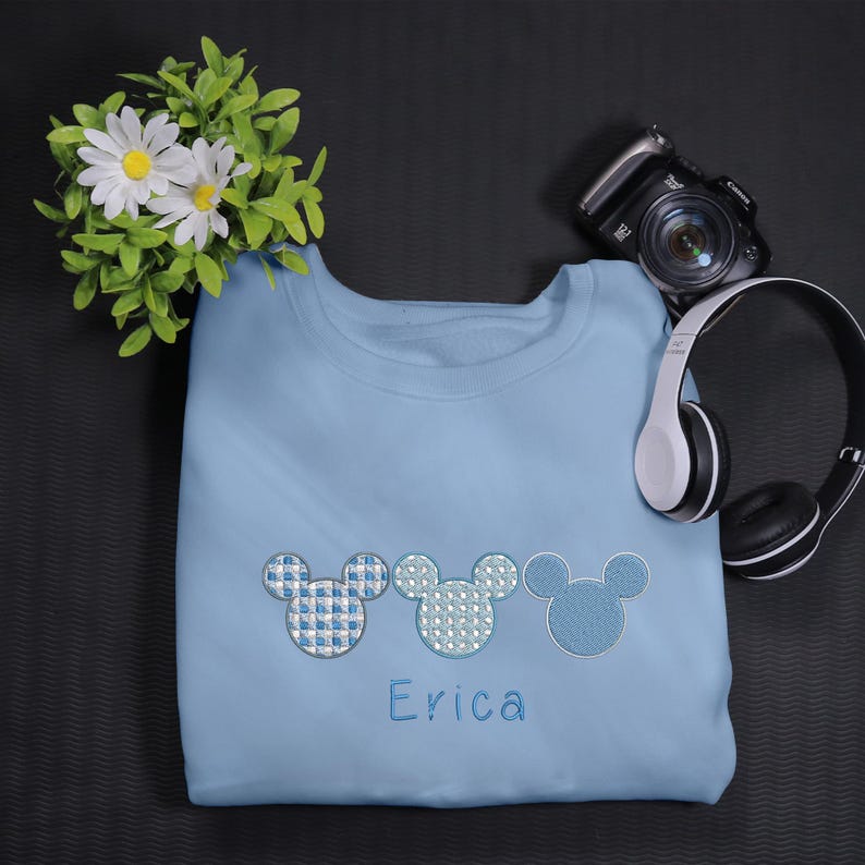 May include: Light blue sweatshirt with three embroidered Mickey Mouse heads in different blue patterns. The name "Erica" is embroidered below the Mickey Mouse heads.