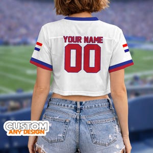 K&ouml;nnte beinhalten: Wei&szlig;es Mesh-Crop-Top-Trikot mit roten und blauen Akzenten. Das Trikot zeigt den Text "YOUR NAME" &uuml;ber der roten Nummer "00". Die &Auml;rmel haben rot-wei&szlig;-blaue Streifen. Der Saum des Trikots ist blau.