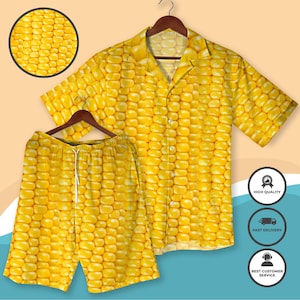 Op de afbeelding: Een geel shirt en short set met maïsthema. De button-down shirt met korte mouwen en de shorts zijn bedekt met een herhalend patroon van gele maïskorrels. De set wordt gepresenteerd op een houten hanger.