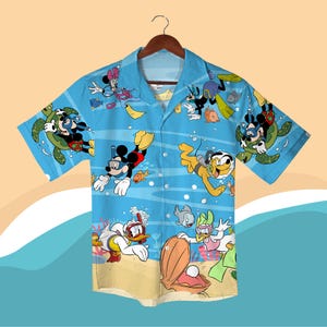 Puede incluir: Camisa de manga corta con botones, con un fondo azul claro que presenta personajes de dibujos animados con equipo de buceo. Mickey Mouse, el Pato Donald y otros personajes de Disney se representan nadando bajo el agua con peces y tortugas. La camisa está colgada de una percha de madera.