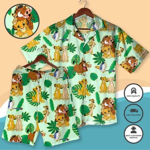 Puede incluir: Un conjunto de camisa y pantalón corto verde claro con un estampado repetido de personajes de El Rey León, incluyendo a Simba, Timón, Pumba y Zazú, junto con hojas tropicales. El conjunto se muestra en una percha de madera. La camisa tiene mangas cortas.